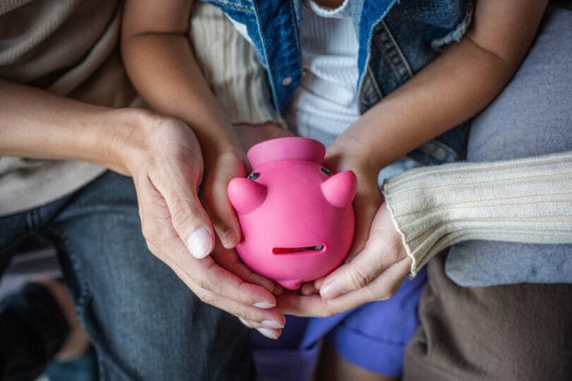 A little girl and her parent holding a pink piggy bank at home. Youth investing, mentored by adult concept