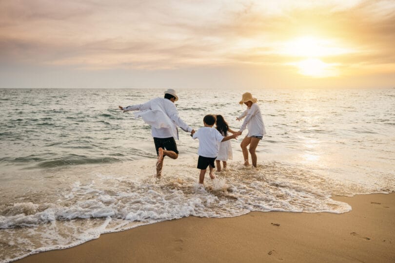Happy Asian parents and daughters have delightful time on beach. Little girls bond with their mother and father running jumping and pretending to fly with arms wide open. Family fun under sun.