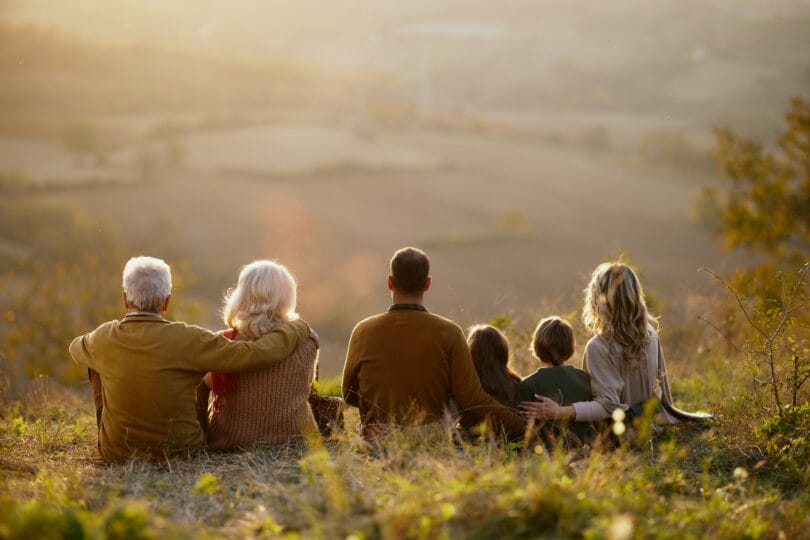 Rear view of carefree multi-generation family looking at view while relaxing in grass during autumn day on a hill. Copy space.