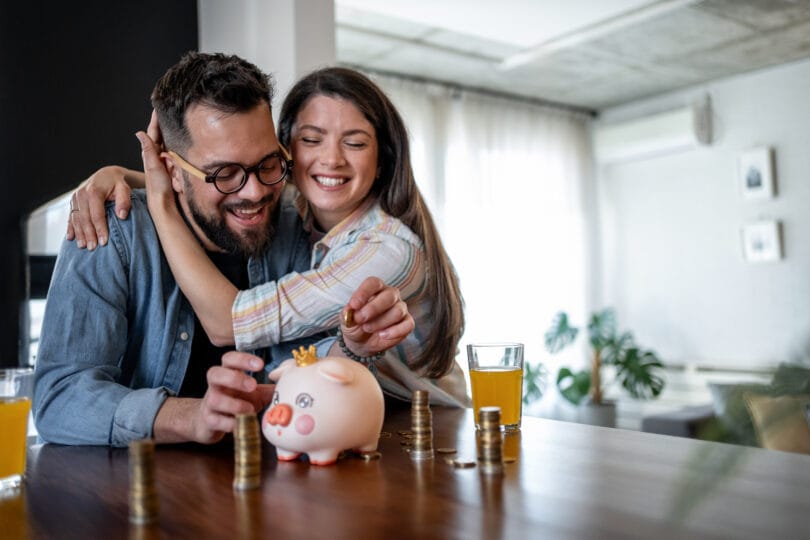 Young, cheerful couple saving money together at home, placing coins into their piggy bank while sharing a warm hug, embodying love and togetherness in their financial journey