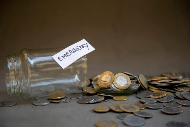 A shattered glass jar labeled Emergency surrounded by scattered currency coins symbolizes financial crisis, depleted savings, and the struggle to manage unexpected expenses and emergencies.