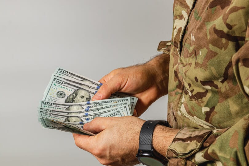 A soldier in camouflage gear counts a stack of cash with focused determination in a neutral-toned indoor environment, highlighting the serious nature of financial matters.