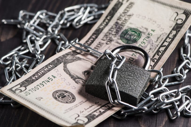 Five dollars are attached with a metal chain and a block. The money is symbolically locked under guard. The composition conveys the idea of financial control.
