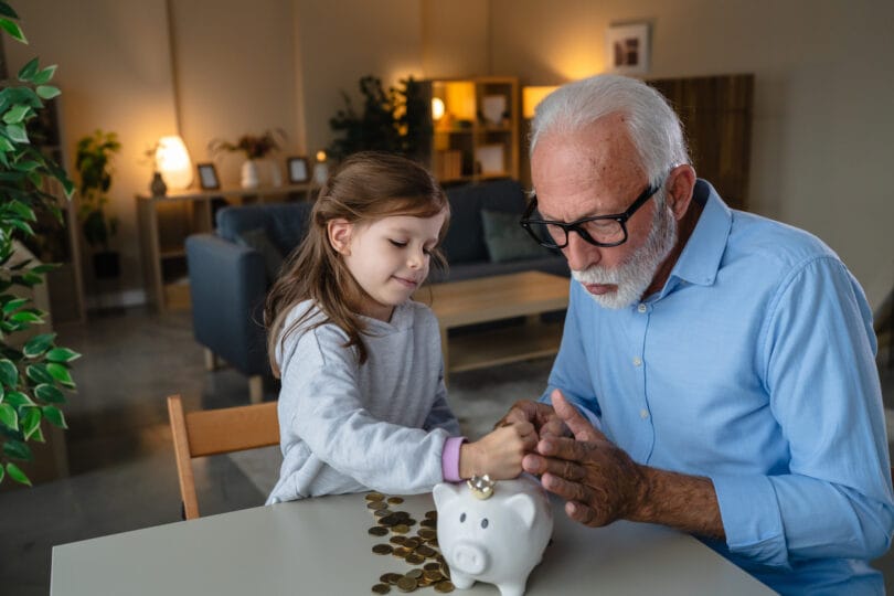 Grandfather teaching his happy granddaughter about financial literacy and the importance of saving money by putting coins into a white piggy bank, creating a strong bond